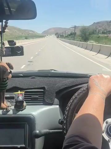 The video is captured from the driver's perspective inside a car traveling on a highway. The dashboard is visible with a fuzzy black mat, a hanging teddy bear, and other small figurines. The driver's hands are on the steering wheel. The road ahead is straight with multiple lanes and a clear blue sky. Mountains and vegetation are visible on the sides of the road. A speed limit sign indicating 90 is visible on the right side of the road. Other cars are seen in the distance. The sun is shining brightly, suggesting a daytime scene.