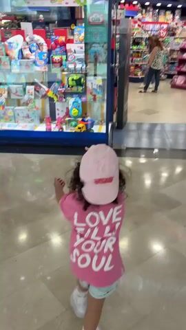 A young child wearing a pink t-shirt with "LOVE FILLS YOUR SOUL" written in white letters on the back and a pink baseball cap walks through a brightly lit toy store. The child is facing away from the camera and is moving towards the back of the store where two women are visible. One woman, wearing a patterned top and jeans, is interacting with another woman, who is wearing a blue apron and holding a doll. The store is filled with shelves of toys and other merchandise. The floor is tiled with a reflective surface. The video appears to be shot from a low angle, following the child as they move through the aisle.