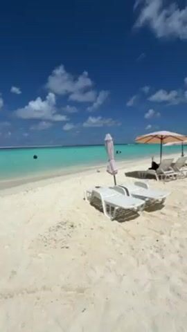 The video shows a tropical beach on a sunny day. The sand is white and the water is turquoise. There are several people swimming in the water, which is calm. There are white plastic beach chairs and umbrellas set up on the sand. The sky is blue with scattered white clouds.