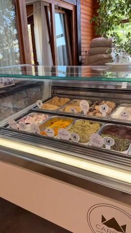 The video pans across a display case filled with various flavors of gelato. The display case is lit from below, illuminating the different colorful options. In the background, there is a rustic wooden interior with plants and outdoor seating visible through windows. A person is seated at a table in the distance, facing away from the camera. The gelato display case has a logo that reads "Carevic" with a crown emblem. Various cups are stacked on the counter behind the gelato. The video also briefly shows a display of what appears to be pastries or other food items in a separate refrigerated case.