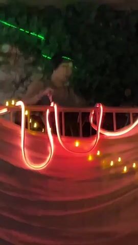 A person with short dark hair is standing behind a DJ setup, illuminated by green laser lights. They are wearing a dark top. The DJ setup is decorated with red and white rope lights and small yellow string lights. A white translucent fabric hangs in front of the setup, further draped with more string lights. The environment appears to be outdoors at night, with greenery visible in the background.