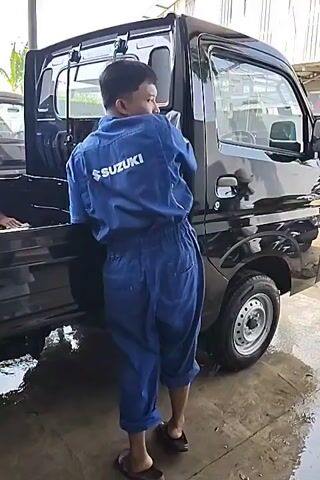 A person wearing a blue Suzuki mechanic jumpsuit is seen washing the side of a black Suzuki truck. The person is using a cloth to wipe down the truck's door and side panel. Water is visible on the ground around the truck, suggesting a car wash setting. Another person can be seen in the background, also near the truck. The lighting indicates it is daytime, possibly indoors in a workshop or under a covered area.