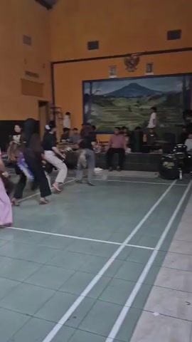 A group of young women in casual clothing are performing a dance in an indoor gymnasium. They are dressed in what appear to be elaborate horse costumes, with riders holding the front part of the costume and a long, flowing tail extending behind them. The dancers are moving in unison, with their bodies and the tails of their costumes creating a fluid, dynamic motion. The gym floor is tiled in a green and white pattern with white lines marking court boundaries. The walls are painted orange, and there is a mural of a landscape scene on the back wall. Spectators are seated along the sides of the room, watching the performance. The lighting appears to be fluorescent overhead.