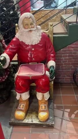 This video shows a festive Christmas display, likely in a store. In the foreground, a life-sized statue of Santa Claus sits in a gilded chair. He is dressed in his traditional red suit, white beard, and a fur-trimmed hat. He holds a small red case on his lap. To the right of Santa, a wooden sleigh is filled with various gift bags. Some bags are plain beige with red ribbons, while others are decorated with Christmas imagery like snowflakes and gnomes. Behind the sleigh, another Santa Claus figure is seated, partially obscured by the gift bags. The background includes a brick wall, a staircase with a green railing, and a Christmas tree. The flooring appears to be terracotta tiles. The lighting suggests an indoor setting.