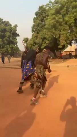 The video shows a group of people dancing on a dirt ground in what appears to be a rural village. Three people are in the foreground dancing.  One of them is wearing a dark shirt and jeans and carrying a stick. Another is wearing a blue and yellow patterned cloth. The third dancer is wearing a leopard print dress.  Other people are watching the dance, and there are trees and buildings in the background. The video is shaky and the lighting is bright.
