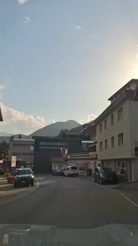 The video shows a street scene in a town with buildings and mountains in the background. The sky is blue with some white clouds. There are several cars parked and driving on the street. One building has a sign that says "El Banca Cantunala". Another building has a sign that says "Volg". The scene appears to be during the daytime.