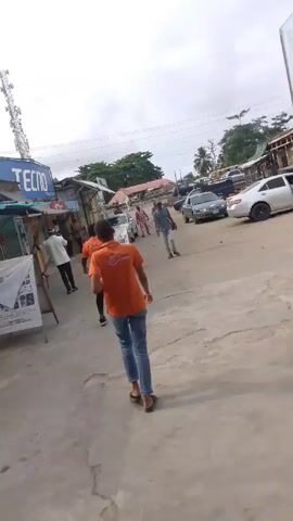 The video depicts a street scene in what appears to be a developing country. Several people are walking along the concrete road, including two individuals wearing orange shirts and jeans. Cars are parked on the side of the road. Buildings line the street, including a shop with a "Tecno" sign and another with "Peace Phones" on its facade. The sky is overcast. A cell tower is visible in the distance. The overall scene suggests a typical day in a busy, somewhat rundown commercial area.