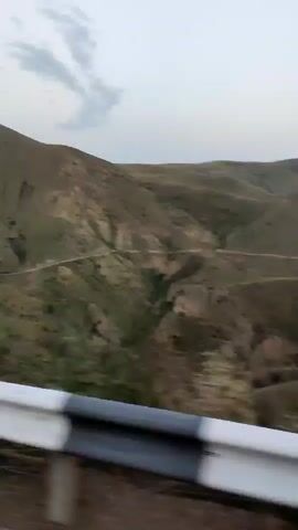 The video shows a panoramic view of a mountainous landscape from a moving vehicle. The terrain is characterized by rolling hills covered in dry grass and sparsely vegetated areas. A winding dirt road snakes its way through the valleys and up the hillsides. In the distance, the slopes of the mountains appear terraced in some areas, suggesting past or present agricultural activity. The sky is overcast with diffused light, creating a muted color palette for the scene. The foreground of the video is dominated by a white barrier with black and white markings, typical of roadside infrastructure. The vegetation in the immediate foreground is blurred due to the motion of the vehicle, creating a sense of speed and movement. There are a few small, brightly colored tents visible on a lower hillside in the left portion of the frame, hinting at a possible campsite or dwelling.