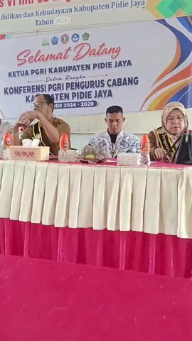The video shows an indoor event with several people seated at a table draped with a pink and white tablecloth. A banner behind them reads "SELAMAT DATANG KETUA PGRI KABUPATEN PIDIE JAYA KONFERENSI PGRI PENGURUS CABANG KABUPATEN PIDIE JAYA PERIODE 2024-2029". There are microphones, bottles of water, and papers on the table. A man in a brown shirt and glasses is speaking into a microphone and gesturing with his hand. Another man in a patterned shirt is seated next to him, and a woman in a headscarf is seated on the other side. In the foreground, a laptop is open on a pink surface. The room appears to be a classroom or meeting hall with windows in the background. The overall atmosphere suggests an official meeting or conference.