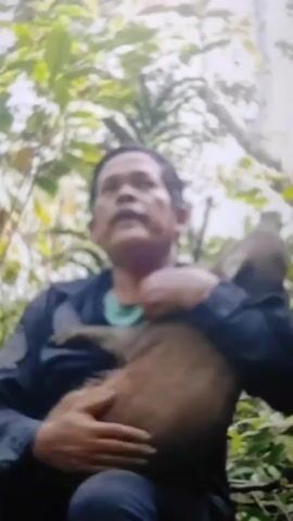 A man is holding a monkey in his arms outdoors. The man is wearing a blue jacket over a light green shirt. The monkey is brown and is resting against the man's chest. The man is looking upwards and appears to be speaking. The background is blurred and consists of green foliage, suggesting a forest or jungle environment.