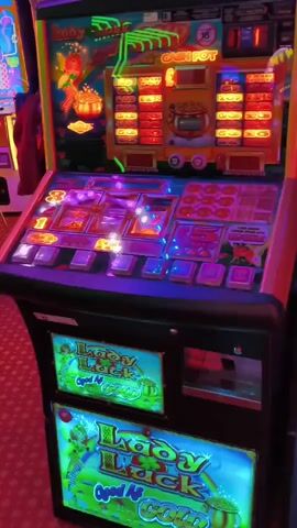 The video shows a close-up, eye-level shot of a "Lady Luck and Gold" slot machine in a dimly lit casino. The machine is black with illuminated, colorful displays on the top, middle, and bottom sections. The top display shows a jackpot amount of 320, with various symbols and paylines. The middle section has spinning reels with colorful symbols, and a control panel with buttons and lights. The bottom section features two identical "Lady Luck and Gold" graphics. The machine is active, with lights flashing and symbols on the reels appearing to spin. The background is blurry but suggests a casino environment with red carpet and other slot machines. No people are visible. The video captures the visual experience of the slot machine in operation.