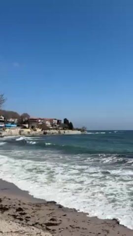 The video shows a beach scene under a clear blue sky. Waves are crashing on the sandy shore. In the distance, there are buildings and vegetation along the coastline. The camera pans slightly to the right, focusing on the sea and the beach.
