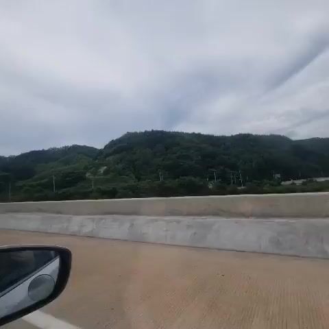 The video appears to be recorded from inside a moving vehicle. The camera is looking forward, with a side mirror visible in the lower left corner reflecting the interior of the car and the road. The road is paved with asphalt and has white lane markings. To the right of the road is a concrete barrier. Beyond the barrier, there is a green, hilly landscape covered in trees. The sky is cloudy and overcast. In the distance, other vehicles can be seen on the road. One black SUV is directly ahead, and a few white cars are further in the distance. The overall impression is of a scenic drive on a highway through a natural environment.