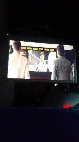 A television screen mounted in a dark vehicle is showing a Formula 1 documentary. Two men in suits are watching the screen. The program transitions from a car race to a man in a suit.