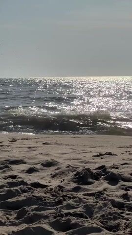 A low-angle, vertical video shows a sandy beach in the foreground and the ocean in the background. The sand is textured with visible grains and small indentations. The ocean is dark blue with bright, shimmering sunlight reflecting off the surface. Small waves are breaking on the shore. The sky is a pale, hazy blue. There are no people or objects visible in the frame. The scene appears to be quiet and still, with only the natural sounds of the ocean.