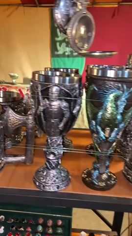 The video shows a collection of decorative mugs and goblets on a shelf. The mugs are metallic with sculpted designs featuring knights, skulls, dragons, wolves, and other fantastical elements. The goblets are similarly decorated. Beneath the shelf is a display case containing rings. The background includes banners or tapestries in varying colors. The scene appears to be a vendor's booth at a market or festival. The camera slowly pans across the display.