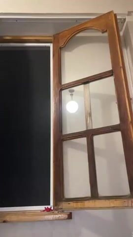 The video shows an interior shot of an open wooden window with multiple glass panes. A black screen is visible behind the window on the left side. In the right panes of the window, a light fixture with a spherical glass shade is reflected, suspended from the ceiling. The light is on and illuminated. The wooden frame of the window appears old and somewhat worn. The walls surrounding the window are a neutral color.