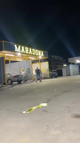 The video shows a nighttime outdoor carwash facility. The camera pans right, revealing the words "MARADONA" and "AUTO SPA H2O IP 71" illuminated in yellow on signage above the washing bays. Several cars are being washed or are parked at the facility. There are several individuals at the location, including a man in shorts and a t-shirt, moving around. The ground is concrete with some puddles or spots visible. The sky is dark with bright lights illuminating the scene.