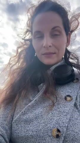 A woman with long brown curly hair and headphones around her neck is looking into the camera. She is wearing a grey sweater with buttons. She is standing outside on a beach with the ocean and cloudy sky in the background.