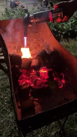 A person is using a propane torch to ignite charcoal in a portable barbecue grill outdoors at night. The grill is dark metal and sits on a stand in the grass. Flames and smoke are visible as the torch is held over the charcoal, which is glowing red. The background is dark and out of focus, with some greenery visible.