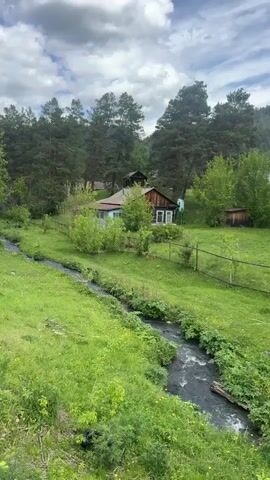 The video shows a natural landscape with a small stream flowing through a green meadow. A modest house is visible in the background, nestled among trees. The sky is cloudy. There are no people immediately visible, but a figure can be spotted on the porch of the house. The water in the stream appears dark. The overall atmosphere is serene.