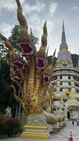 This video shows the exterior of a Buddhist temple. A large, multi-headed golden dragon statue is in the foreground on the left. Behind the statue is a multi-tiered, ornate temple building with a tall spire. Several smaller spires are also visible. People are walking on a paved area in front of the temple. The environment appears to be a tropical or sub-tropical setting with lush green mountains in the background. The sky is overcast with some clouds. The overall impression is one of grandeur and religious significance.