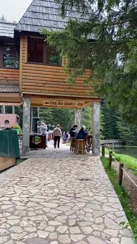 The video shows an outdoor scene in Pitomine, Montenegro, featuring a rustic wooden building with a stone foundation and pillars. A stone path leads under an archway formed by the building. Above the archway, a wooden sign with an arrow points left and reads "Vine and souvenir shop."

Several people are present. Some individuals are walking on the stone path, moving away from the camera and under the archway. To the right, under the archway and next to what appears to be a lake or pond, a few people are sitting at wooden tables on high stools, seemingly enjoying the outdoor setting. To the left, under the archway, another group of people is standing near a display or counter. The building itself has multiple windows on its upper floor, reflecting the green surroundings. Lush green trees and a forest are visible in the background, further emphasizing the natural setting. The weather appears bright and clear, indicative of daytime.