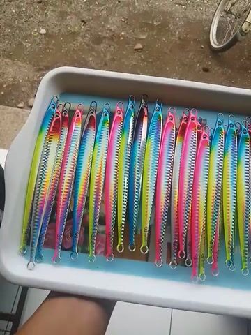 Here is a concise description of the video:

The video shows a white plastic tray filled with about 20 fishing lures. Each lure is long and slender, with a colorful, rainbow-like pattern consisting of pink, yellow, blue, and green stripes. The lures have a metal loop at each end for attaching fishing line.  The tray appears to be sitting on a concrete surface, with a bicycle tire partially visible in the upper left corner. The camera moves slightly around the tray, offering different perspectives of the fishing lures.