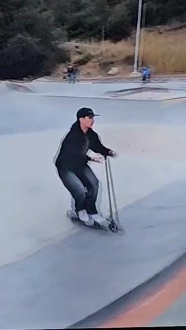 A person dressed in dark clothing is performing a flip on a scooter at a skate park. The skate park has concrete ramps and bowls, with trees and a road visible in the background. A white car is seen driving on the road. A person on a bicycle is also visible in the background.