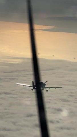 This video shows a small propeller airplane flying above a layer of clouds during sunrise or sunset. The sun is low on the horizon, casting a golden light on the clouds. Several other aircraft are visible in the distance, also flying above the clouds. The camera appears to be mounted inside another aircraft, with a portion of its wing or strut visible in the foreground, slightly out of focus.