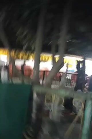 The video shows a dark outdoor scene at night, likely a market or food stall area. In the foreground, there are large tree branches with dark leaves obscuring the view. Behind the branches, a yellow awning is visible, under which several large, reddish-orange round objects, possibly food containers or cooking pots, are arranged in a line. There are several people in the background, some standing near the awning and others appearing to be walking. A black railing or fence with a decorative gold design is also visible in the lower part of the frame. The lighting suggests artificial illumination, casting shadows and creating a dim atmosphere.