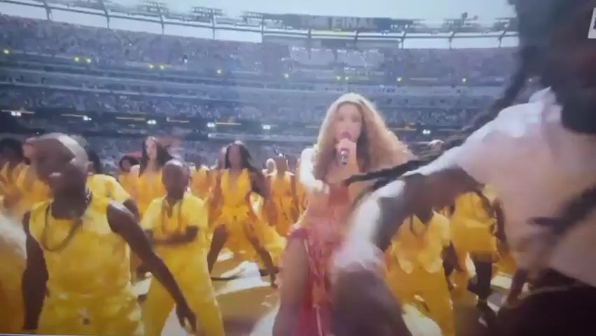 This is a screenshot from a video showing a live musical performance on a large stadium stage. A woman with long blonde hair, wearing a pink and yellow sequined outfit with a high slit, is singing into a pink microphone. She is positioned in the center of the frame and appears to be dancing. She is surrounded by a large group of performers, many of whom are children, all wearing matching bright yellow outfits. The background shows rows of stadium seating filled with spectators. The overall lighting suggests daytime or a well-lit stadium setting. The word "THE FINAL" is visible on a banner in the upper part of the stadium seating.