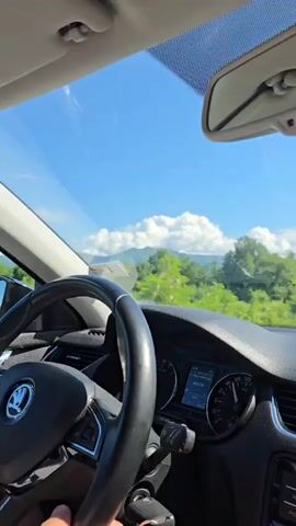 The video shows a car driving on a highway on a clear sunny day. From the driver's perspective, the car is on a straight stretch of road, with trees and vegetation lining both sides. In the distance, mountains can be seen against the blue sky with scattered clouds. The dashboard is visible, with the steering wheel, instrument panel, and central console in view. A small air freshener hangs from the rearview mirror. There are a couple of other cars on the road ahead.