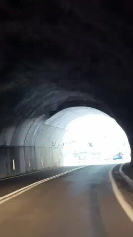 The video shows a view from a car driving through a mountain tunnel. The tunnel is made of concrete and rock. As the car exits the tunnel, it reveals a scenic mountain landscape with a winding road. Several cars are seen driving on the road, and a few people are standing on the side of the road. The camera pans to the right, showing a black car parked on the side of the road with a couple standing next to it. The man is wearing a dark shirt and pants, and the woman is wearing a light-colored dress. They appear to be taking photos or enjoying the view. The video ends with a close-up of the couple and the parked car.