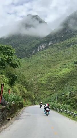The video shows a group of people riding motorbikes on a paved road winding through a lush, green mountainous landscape. The mountains are partially obscured by low-hanging clouds, creating a misty atmosphere. The roadside is lined with vegetation, including tall corn stalks. Streetlights are visible along the side of the road. The sky is overcast, suggesting a cloudy day.