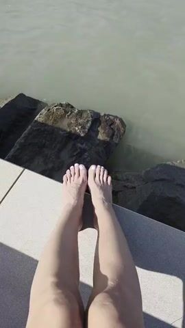The video begins with a close-up shot of a pair of bare feet with white painted toenails resting on a light-colored surface, possibly a dock or walkway. Beyond the feet, large, dark rocks are visible, partially submerged in calm, murky water. The camera then slowly pans upwards, revealing more of the water and the surrounding environment. Several boats are anchored in the distance on the water. One boat, a sailboat, is prominent in the frame. Other boats, some covered, are also visible. The horizon shows a distant shoreline with buildings and vegetation. The sky appears overcast with patches of blue. The video ends by focusing on the boats and the expanse of water.
