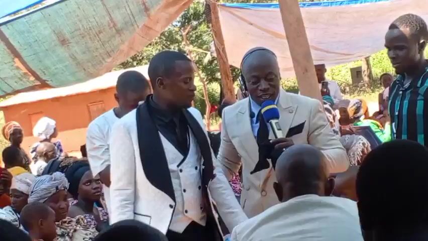 Certainly. Here is a description of the video:

The video shows a group of people gathered outdoors, likely in a rural or semi-rural setting. A man dressed in a light-colored suit and tie is holding a microphone and speaking to another man who is wearing a white jacket. Several other people are standing around them, including one who is recording the scene with a smartphone. The setting appears to be under a makeshift shelter with a white tarp supported by wooden posts. There are trees and possibly some simple structures visible in the background. The man speaking into the microphone seems to be addressing the crowd, possibly in a formal or religious context.