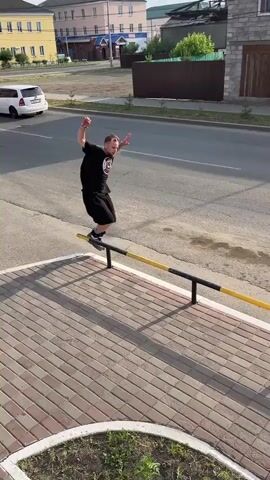 A young adult male wearing a black t-shirt, black shorts, and rollerblades is performing a trick on a black metal rail with yellow markings. He is balancing on the rail on his rollerblades, with his arms outstretched for balance. The rail is positioned alongside a red brick sidewalk. In the background, there is a street with cars, buildings, and trees. Another person can be seen in the distance on the street, also on rollerblades. The scene appears to be an urban or suburban environment during the daytime.
