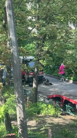The video shows a street sweeper positioned on a paved road next to a red trailer. A woman in a pink jacket walks away from the sweeper, down the road and out of view. The sweeper is red and gray, with rotating brushes at the front. It is parked next to a section of green metal fencing and a patch of green vegetation. The scene is partially obscured by a tree with green leaves and a gray utility pole. A blue car is also partially visible behind the tree.