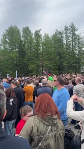 Here is a description of the video. 

The video shows a crowd of people at an outdoor event. They are facing a stage with a banner that reads "С ДНЁМ ХИМИКА!", which translates to "Happy Chemist's Day!". Many in the crowd have their hands raised in the air. In the background are green trees and an overcast sky.