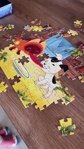 A young blonde girl wearing a green t-shirt with a cat print is sitting at a wooden table, assembling a jigsaw puzzle. The puzzle depicts a cartoon dog with a long tongue reaching towards an orange cartoon character. Scattered puzzle pieces are visible on the table and the wooden floor. The girl is focused on the puzzle, and her mouth is slightly open. She then turns and walks away from the table. The environment appears to be a living room or dining area with wooden flooring and walls.