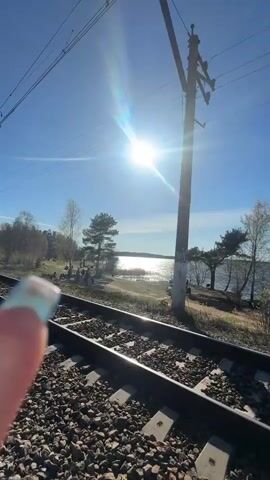 The video captures a scenic outdoor setting in Toksovo, Russia, on a bright, sunny day. The clear blue sky is prominent throughout, with a strong sun flare visible in the initial frames.

The primary subject is a set of railroad tracks, visible in the foreground with gravel ballast between the rails. To the left of the tracks, a large body of water—likely a lake or wide river—stretches into the distance. Several small groups of people are gathered on the grassy shore beside the water, possibly enjoying the weather or engaged in activities like fishing. The trees along the shore appear mostly bare or sparsely leaved, suggesting early spring or late autumn. Overhead electricity lines and poles run alongside the tracks.

Around the 0:04 mark, the perspective shifts to follow a person walking away from the camera along a dirt path next to the railroad tracks. This individual is wearing a black hoodie and a black backpack, on which the brand name "PUMA" is clearly visible. They are carrying two plastic bags, one white and one orange. Other people are also seen walking further down the tracks in the distance. To the right of the tracks in the background, a building structure is partially visible. The overall atmosphere is tranquil and pleasant under the bright sunshine.