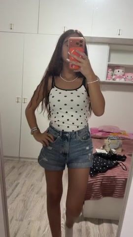 The video shows a young woman taking a mirror selfie in a bedroom. She is wearing a white polka dot tank top, denim shorts, and white socks. She has long brown hair and is holding a phone to take the picture. She poses with her hands on her hips and then runs her hand through her hair. The room has a large white wardrobe, a bed with pink and white bedding, and a wooden floor. There are shelves in the wardrobe with stuffed animals.