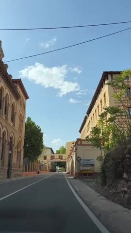 The video captures a view from a moving vehicle, likely a car, traveling down a road lined with buildings. On the left, a tan-colored building with arched windows and a tiled roof is visible. On the right, a larger, yellow building with red trim, multiple windows, and balconies dominates the scene. Trees are present along the left side of the road, and there is an archway ahead with the text TERMAS PALLARES. A sign with traffic symbols, including a height restriction and directions to Alhama, is posted on the right side of the road. The sky is blue with scattered white clouds. The road has lane markings. A single person is visible walking on the sidewalk on the left side of the road.