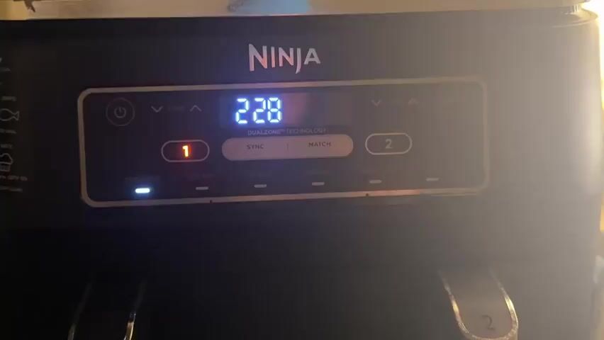 A close-up shot of the control panel of a black Ninja air fryer is shown. The digital display shows the temperature decreasing from 227 degrees to 223 degrees Fahrenheit. The buttons below the display show options for "MAX CRISP", "AIR FRY", "ROAST", "REHEAT", and "DEHYDRATE". The buttons for zones 1 and 2 are visible, as are the "SYNC" and "MATCH" options. The overall lighting is dim and a bright light source is entering the frame from the right, creating glare on the control panel.