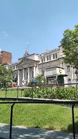 The video presents a wide, static shot of an urban scene on a sunny day. A large, white building with classical architectural features dominates the background, adorned with a flagpole flying a light blue and white flag. In front of the building is a landscaped area with green grass, interspersed with plants. A black metal railing runs along the edge of the landscaping, separating it from a paved walkway. 
To the left of the main building are several high-rise buildings, some with balconies and others with brick facades. Trees are interspersed among the buildings, adding patches of greenery. A small vendor stall can be seen to the right of the main building. The sky is blue with a few scattered white clouds.