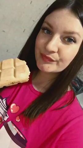 The video features a young woman with long, dark hair and blue eyes, wearing a vibrant pink t-shirt adorned with a graphic featuring a white cupcake or coffee cup and red hearts. She is holding a large, rectangular, light-colored food item resembling a waffle or a thick biscuit, molded into a grid pattern similar to a chocolate bar.

The woman brings the treat to her mouth and takes a large bite, accompanied by a distinct crunching sound. After taking a bite, a darker brown filling is visible inside the food item, suggesting it might be a filled waffle or a sandwich biscuit. She continues to chew with a content and slightly playful expression. Her fingernails are painted with a light color and feature subtle, swirling designs. The setting appears to be indoors, with a plain, light-colored wall with a subtle textured pattern visible in the background. The video is a close-up shot focusing primarily on her face and the food she is eating. No specific time of day or weather cues are evident.