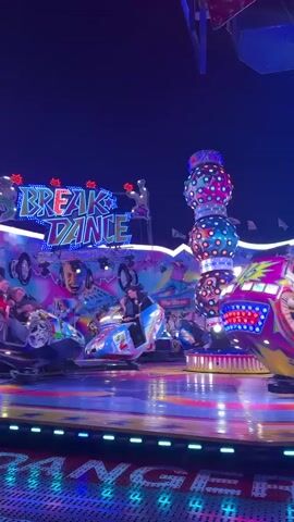 The video shows a breakdance amusement park ride in operation at night. The ride is brightly lit with colorful lights. Several individual cars on the ride, each designed to hold a few passengers, are spinning and rotating. The ride appears to be in a fairground or amusement park setting, with a dark sky in the background. The name "BREAK DANCE" is prominently displayed on a sign on the ride, illuminated with neon lights. People are visible inside the cars, appearing to be enjoying the ride. The cars are decorated with various designs, including one with an American flag motif and a statue of liberty image.