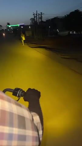 A first-person perspective video shows a person riding a motorcycle down a cracked asphalt road at dusk. The headlight casts a bright yellow glow illuminating the road ahead. On the left, a sign with green and white text is partially visible. Across the street, lights from businesses or homes add color to the dusky background. The sky transitions from dark blue at the top of the frame to a lighter hue near the horizon. Overhead, power lines stretch across the sky. The rider wears a plaid shirt, and their gloved hand is visible on the motorcycle handle.