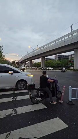 The video shows a street scene with a pedestrian crossing in the foreground. A person is sitting on a scooter, facing away from the camera, at the edge of the pedestrian crossing. To the left of the scooter, a white SUV is passing through the intersection. Further in the background, there is an elevated roadway with traffic. The sky is overcast, suggesting it may be late afternoon or evening. Other vehicles are visible in the distance, and streetlights are on.